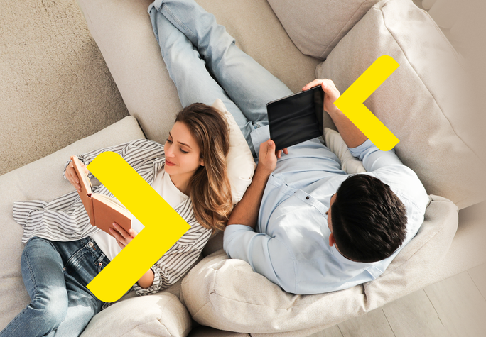 The image shows a couple relaxing on a comfortable sofa. One person is reading a book, while the other is holding a tablet, indicating a shared moment of leisure and engagement with different activities. The cozy setting is enhanced by the soft textures of the sofa and the warm lighting. Yellow arrows are placed on either side of the image, pointing left and right.