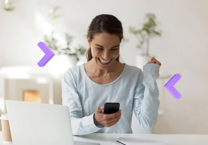  Kobieta w białej bluzie siedzi przy biurku, trzymając smartfon w jednej ręce, a druga ręka jest uniesiona w geście entuzjazmu. Na biurku znajduje się laptop, a w tle są rośliny w białych doniczkach.