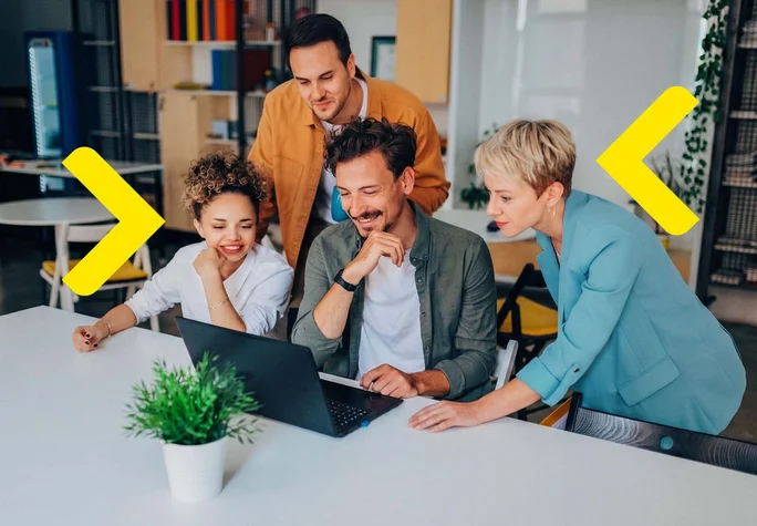 The image shows a group of individuals collaborating at a table in an office. They are focused on a laptop, analyzing data or a project. The environment is modern, featuring colorful furniture and plants, which creates a friendly atmosphere that fosters creativity and collaboration. The participants are dressed in a variety of stylish outfits, adding energy to the scene.