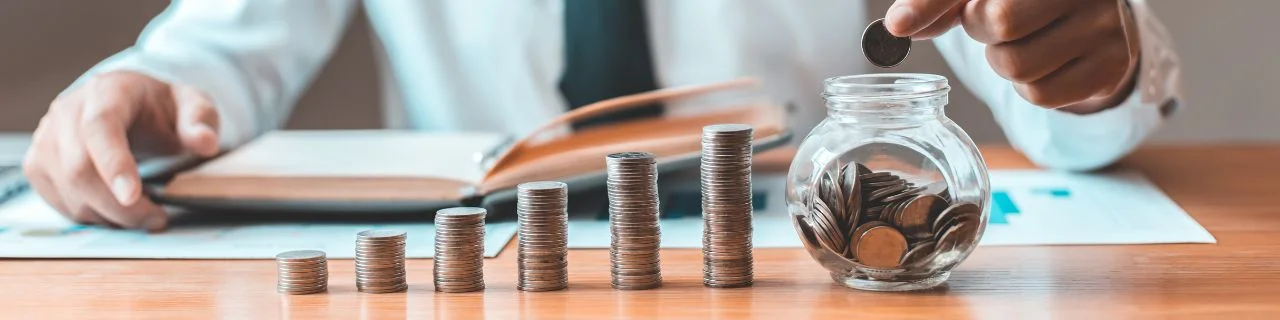 Ręka wkładająca monetę do słoika, symbolizująca oszczędzanie i zarządzanie finansami, na tle dokumentów i książki na biurku.