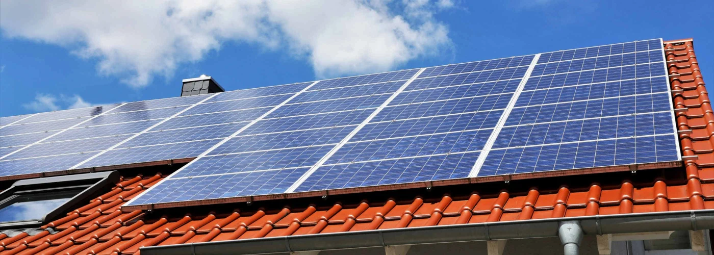 Zdjęcie przedstawia nowoczesną instalację paneli słonecznych zamontowanych na czerwonym dachu budynku. Tło stanowi jasne niebo z kilkoma chmurami, co podkreśla ekologiczne i energooszczędne rozwiązania w zakresie odnawialnych źródeł energii.