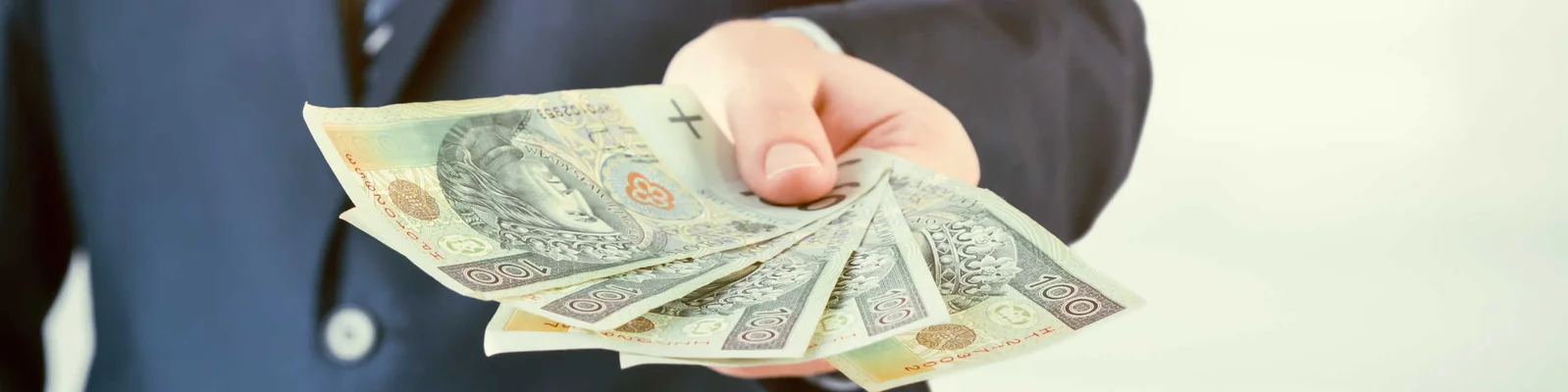 Mężczyzna w garniturze trzymający w ręku plik rozłożonych w wachlarz banknotów o nominale 100 zł