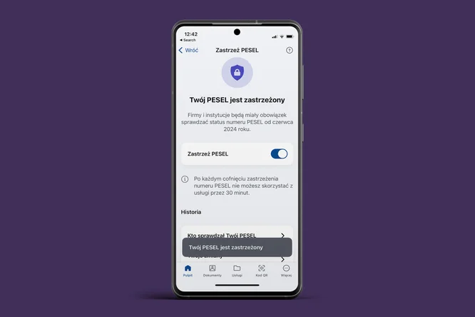 Na zdjęciu widoczny jest telefon komórkowy z białym wyświetlaczem na tle fioletowym. Ekran pokazuje interfejs aplikacji dotyczącej zarządzania numerem PESEL. U góry widnieje ikona z tarczą oraz napis "Zastrzeż PESEL". Informacja na ekranie wskazuje, że PESEL jest zastrzeżony, a poniżej znajduje się przycisk umożliwiający zastrzeżenie numeru. W sekcji "Historia" widoczna jest informacja o statusie PESEL oraz szczegóły dotyczące ostatnich działań. Na dole ekranu znajdują się ikony reprezentujące różne funkcje aplikacji.