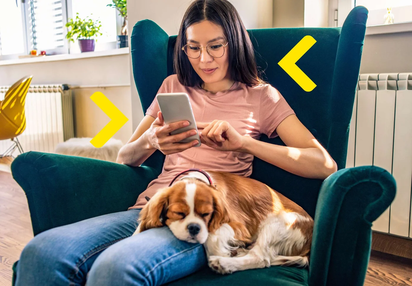 Kobieta w okularach siedzi na fotelu trzyma w ręku telefon a obok śpi pies opierając głowę o jej nogi.