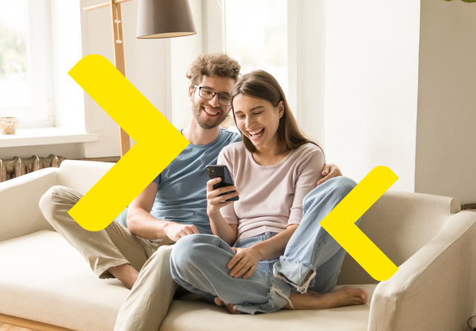 The photo features a couple sitting on a light-colored sofa in a cozy interior. One person is holding a mobile phone, and both individuals are close together, suggesting intimacy and shared time. In the background, there are large windows allowing natural light to flood the space, adding warmth and a pleasant atmosphere. Yellow arrows are placed on either side of the image, pointing left and right.