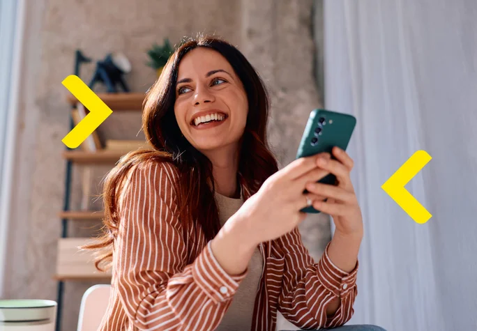 Kobieta siedzi w salonie i uśmiecha się do kogoś z poza kadru, trzymając telefon w rękach
