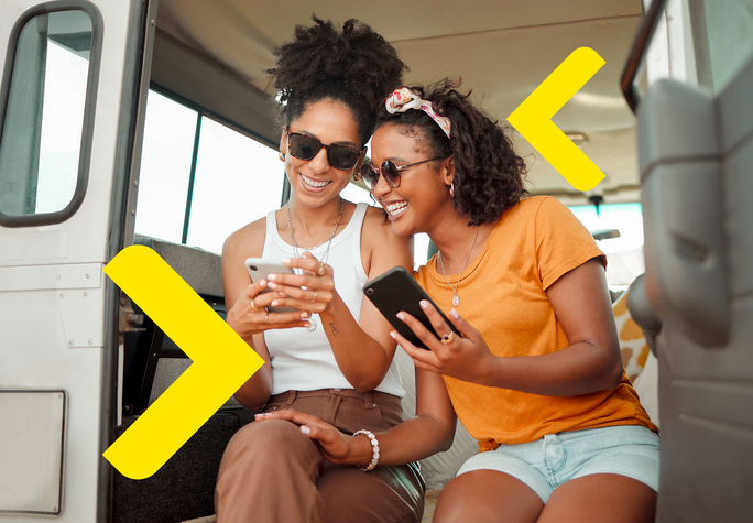 The image shows two friends sitting together in a cozy setting, likely inside a vehicle. Both are engaged with their smartphones, sharing something on their screens, which indicates a moment of connection and enjoyment. The warm colors of their clothing and the inviting atmosphere contribute to a sense of camaraderie. Yellow arrows are placed on either side of the image, pointing left and right.