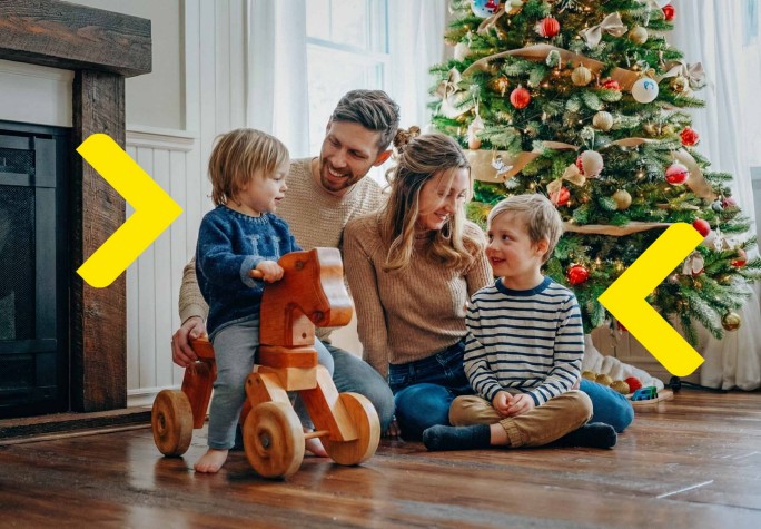 Czteroosobowa rodzina siedzi na ziemi i spędza wspólnie czas. Najmłodsze dziecko siedzi na koniu na biegunach. W tle widać kominek i choinkę