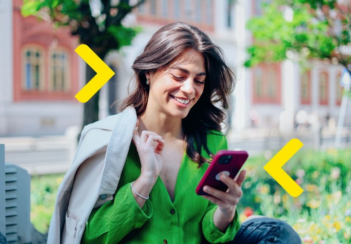 Kobieta siedzi na ławce przed budynkiem wyglądającym jak uczelnia. Patrzy w telefon i ma płaszcz przewieszony na ramieniu