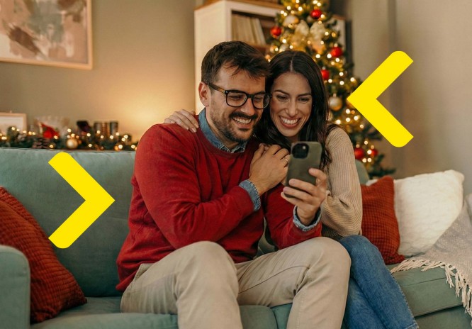 Para siedzi na kanapie i oboje uśmiechają się do telefonu. W tle widać choinkę i inne ozdoby świąteczne