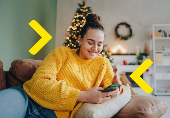 Kobieta siedząc na kanapie relaksuje się korzystając z telefonu i uśmiechając się do niego. W tle widać ozdoby świąteczne, w tym choinkę.