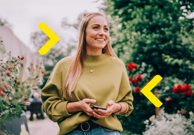 Kobieta stoi na tarasie na zielonym obszarze z drzewami i kwiatami w tle. Patrzy gdzieś w dal i śmieje się, trzymając telefon w rękach. 