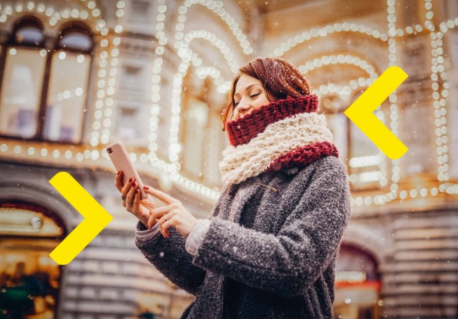 Kobieta ciepło ubrana korzysta z telefonu, stojąc pod oświetloną kamienicą. W tle widać padający śnieg