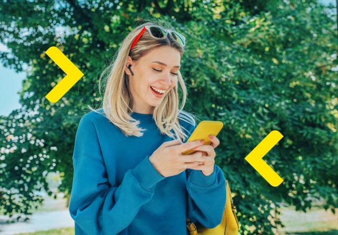 Kobieta przy słonecznej pogodzie jest na spacerze w parku i patrzy w telefon, do którego się uśmiecha. W tle widać drzewa.