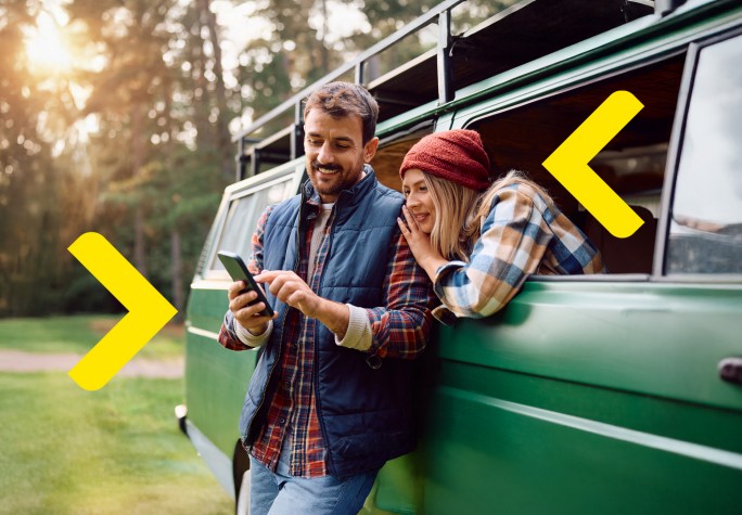 Para ogląda razem coś w telefonie. Mężczyzna stoi oparty o samochód typu kamper. Kobieta siedzi w samochodzie i przez okno zagląda mężczyźnie przez ramię w telefon. Samochód stoi zaparkowany na łące, a w tle widać drzewa.