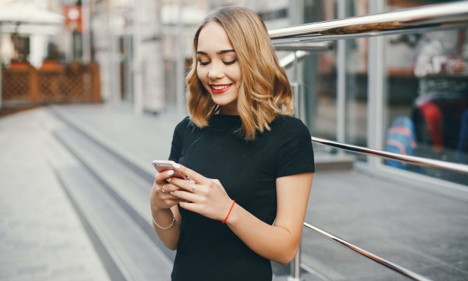 Zdjęcie przedstawia kobietę stojącą na zewnątrz, trzymającą w rękach smartfon. Ma blond, falujące włosy i ubrana jest w czarną koszulkę. W tle widać elementy miejskiej architektury, takie jak szklane ściany i metalowe barierki. 