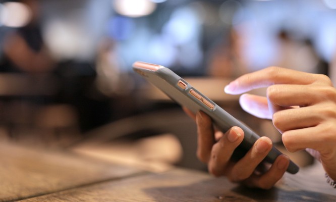 Zdjęcie przedstawia rękę trzymającą smartfon, na którym osoba korzysta z aplikacji lub przegląda treści. W tle widoczna jest przestrzeń kawiarni z rozmytymi postaciami i światłem, co wskazuje na przyjazne i dostępne środowisko publiczne. 