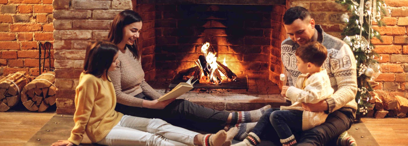 Happy european family with children sitting on floor by fireplace reading and frying marshmallow over fire, young loving parents with two little kids enjoying winter holidays in cozy country house