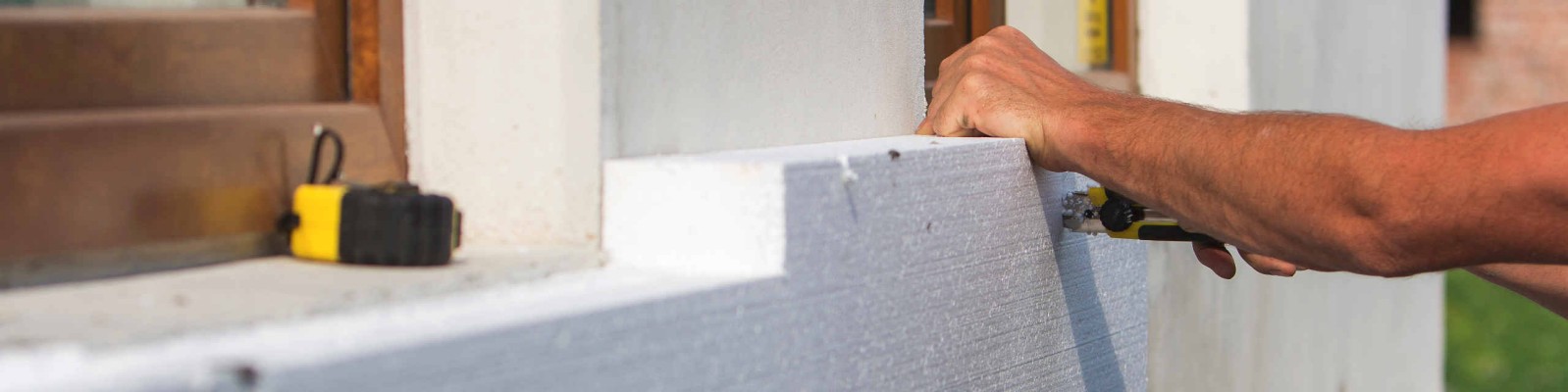 Worker hand with level and knife measuring and cutting white rigid polyurethane foam sheet on wall at newly installed plastic window. Modern technology, renovation, insulation concept.