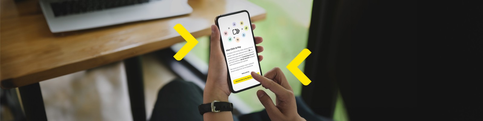Na zdjęciu widać osobę trzymającą smartfon z otwartą aplikacją bankową. Użytkownik korzysta z funkcji płatności, a na ekranie wyświetlają się różne ikony i opcje. W tle znajduje się laptop oraz drewniany stół, co sugeruje, że osoba pracuje w komfortowym otoczeniu. Żółte strzałki wskazują na interaktywność aplikacji, podkreślając łatwość nawigacji i dostępność funkcji.