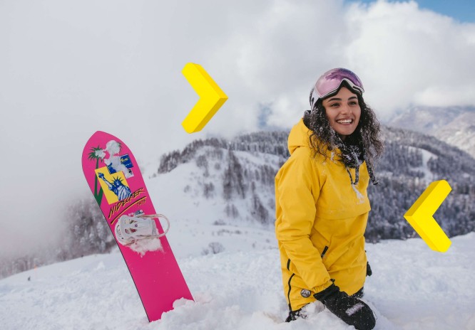 Dziewczyna ubrana w żółtą kurtkę, z googlami na głowie stoi w śniegu obok różowej deski snowboardowej. W tle widać ośnieżone góry.