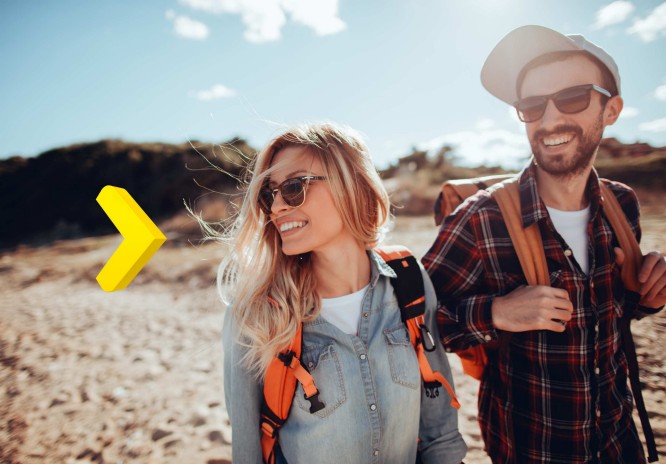 Na zdjęciu uśmiechnięta kobieta z długimi blond włosami i mężczyzna z brodą spacerują po piaszczystej plaży. Oboje mają na sobie okulary przeciwsłoneczne, plecaki turystyczne i luźne, wygodne ubrania – kobieta w dżinsowej koszuli, mężczyzna w koszuli w kratę i czapce z daszkiem. W tle widać wydmy i skąpane w słońcu niebo. Po lewej stronie dodano żółtą strzałkę.