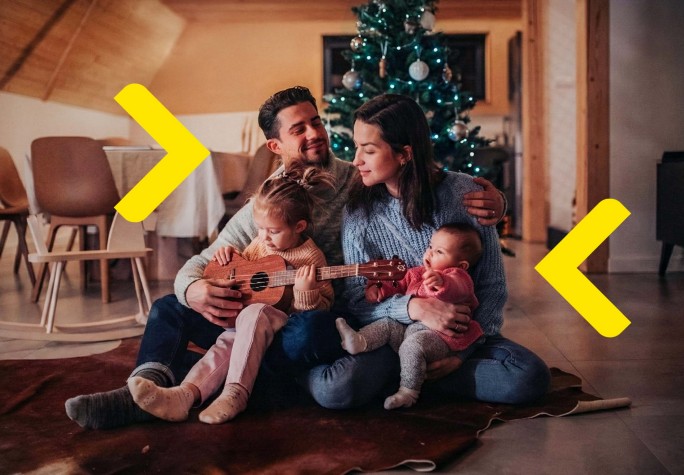 Czteroosobowa rodzina siedzi na podłodze w salonie, przytulając się. Dziewczynka ma gitarę na kolanach. W tle widać choinkę.