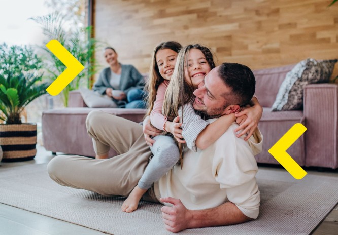 Czteroosobowa rodzina siedzi w salonie. Na pierwszym planie ojciec leży na podłodze, a na nim dwie córki. Wszyscy się przytulaja i śmieją. W tle matka patrzy na całą scenę i też się śmieje