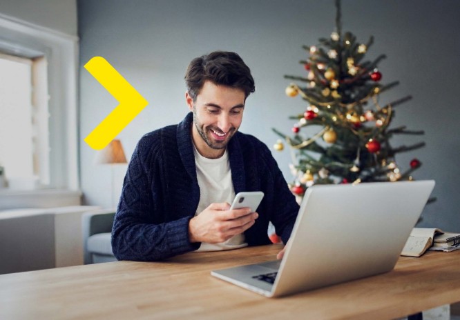 Mężczyzna siedzi przy otwartym laptopie przy stoliku. Korzysta w tym samym czasie z telefonu, uśmiechając się do niego. W tle widać choinkę.