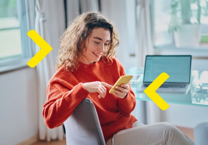 Kobieta siedzi na krześle i korzysta z telefonu z uśmiechem na ustach. Znajduje się w mieszkaniu, siedzi przy biurku odwrócona do niego bokiem. Na stole znajduje się laptop