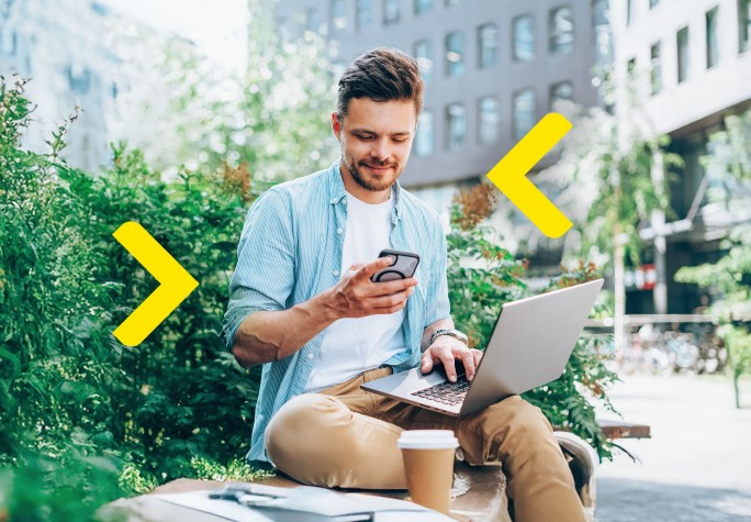 Mężczyzna siedzi na murku przed blokiem, z laptopem na kolanach i kawą leżącą obok. W rękach trzyma telefon, na który się patrzy