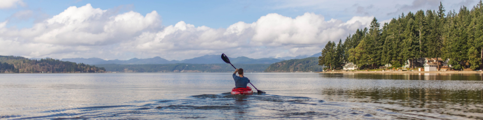Zdjęcie przedstawia osobę w kajaku na spokojnym jeziorze, z widokiem na lasy i góry w tle