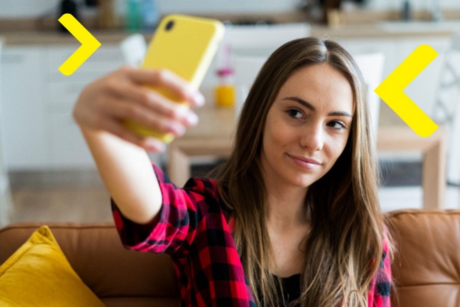 Osoba siedzi na kanapie, trzymając w ręku żółty smartfon i robiąc selfie. Ubrana jest w czarno-czerwoną koszulę w kratę. W tle widać jasne, nowoczesne wnętrze z meblami i dekoracjami.