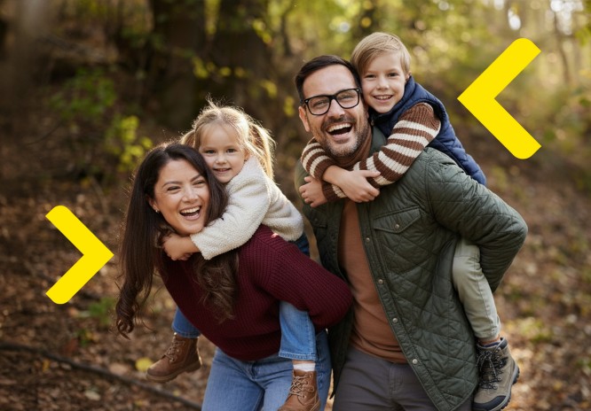 Czteroosobowa rodzina jest na spacerze w lesie w jesiennej aurze. Rodzice trzymają swoje dzieci na plecach. Wszyscy się śmieją.