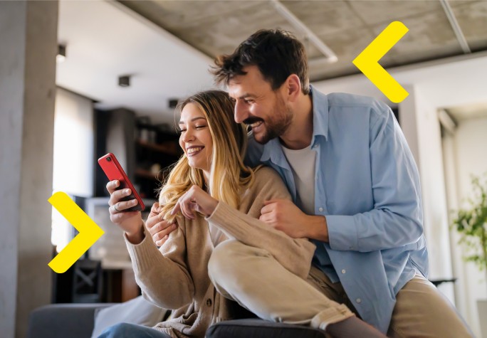 Para siedząc na kanapie obejmuje się oraz ogląda coś na telefonie, śmiejąc się