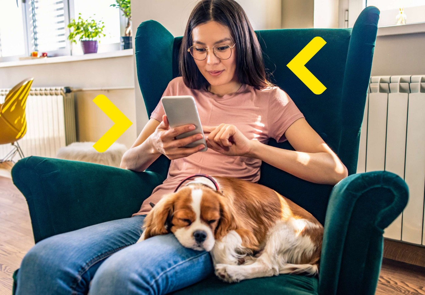 Kobieta w okularach siedzi na fotelu trzyma w ręku telefon a obok śpi pies opierając głowę o jej nogi.