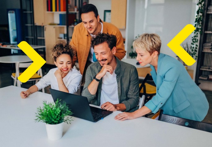 Das Bild zeigt eine Gruppe von Personen, die an einem Tisch in einem Büro zusammenarbeiten. Sie konzentrieren sich auf einen Laptop und analysieren Daten oder ein Projekt. Die Umgebung ist modern, mit bunten Möbeln und Pflanzen, was eine freundliche Atmosphäre schafft, die Kreativität und Zusammenarbeit fördert. Die Teilnehmer sind in verschiedenen stilvollen Outfits gekleidet, was der Szene Energie verleiht.