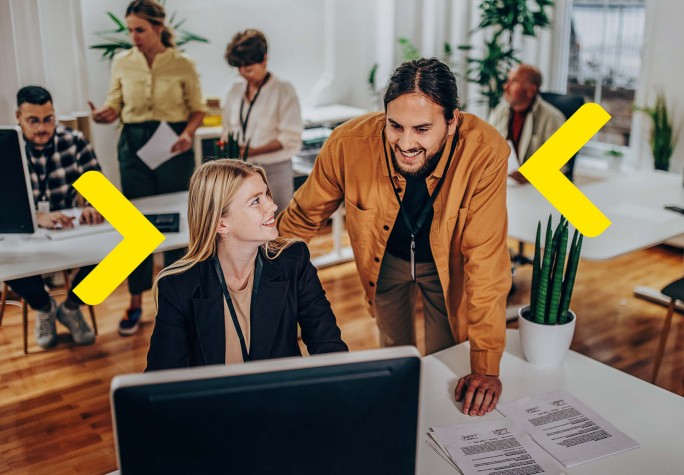 Das Bild zeigt eine Gruppe von Personen, die in einem Büro arbeiten. Der Fokus liegt auf einer Frau, die an einem Schreibtisch mit einem Computer sitzt, während ein Mann neben ihr steht und etwas auf dem Bildschirm bespricht. Im Hintergrund sind andere Mitarbeiter mit verschiedenen Aufgaben beschäftigt. Das Büro ist modern, mit viel natürlichem Licht und Pflanzen, was eine freundliche Arbeitsatmosphäre schafft.