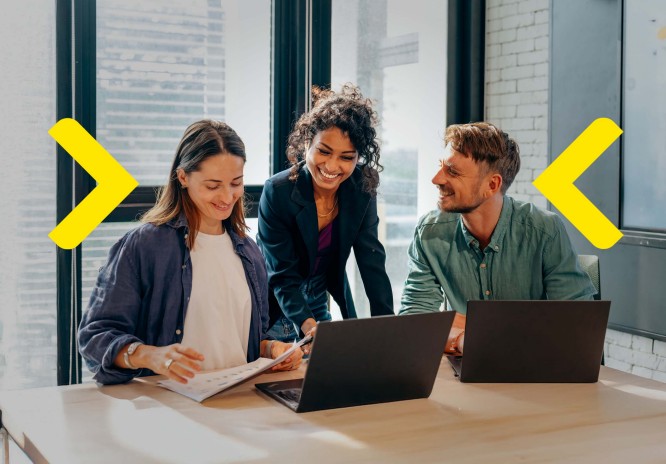 Das Bild zeigt eine Gruppe von Personen, die an einem Tisch in einem modernen Büro zusammenarbeiten. Zwei Personen sitzen vor Laptops und überprüfen Dokumente, während eine dritte Person neben ihnen steht und auf etwas auf einem Blatt Papier zeigt. Große Fenster im Hintergrund lassen viel natürliches Licht herein, was eine helle und inspirierende Atmosphäre schafft, die die Teamarbeit fördert.